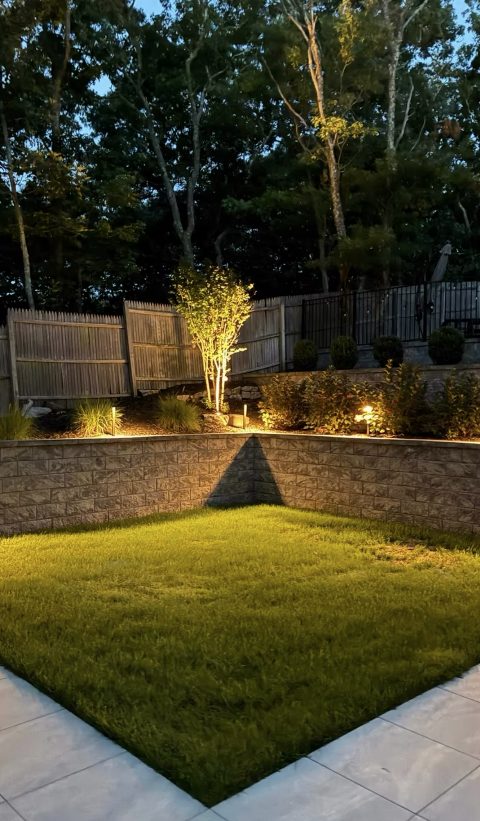 A lit retaining wall, uplit tree and privacy-fenced back lawn on a Long Island property at night